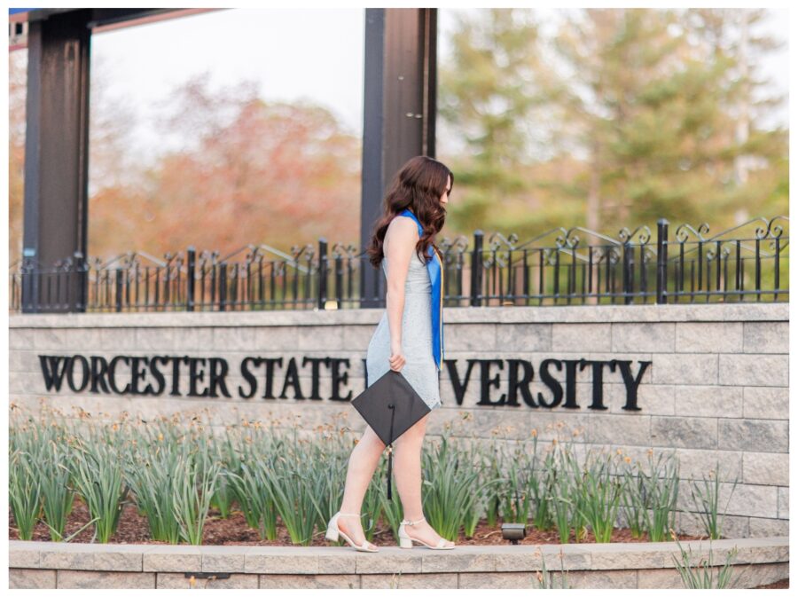 Senior girl holding mortar board walking in front of Worcester State University sign