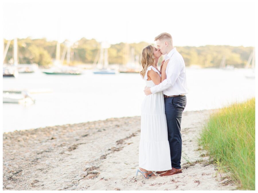 A couple kissing on the beach with boats in the background at The Knob Engagement Session Falmouth, MA Cape Cod