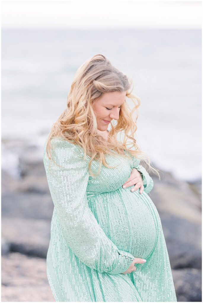 Woman standing on rocks at Old Silver Beach in Falmouth, MA Cape Cod while wearing a green sequined dress and holding baby bump