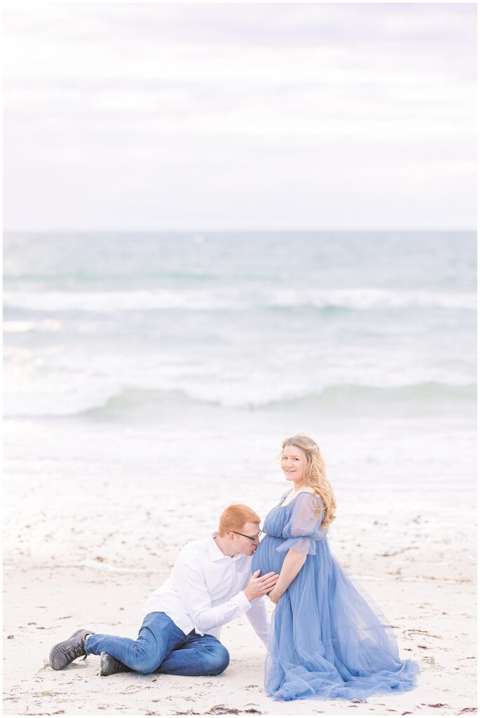 Man sitting on the beach and kissing his wife's baby bump maternity photos