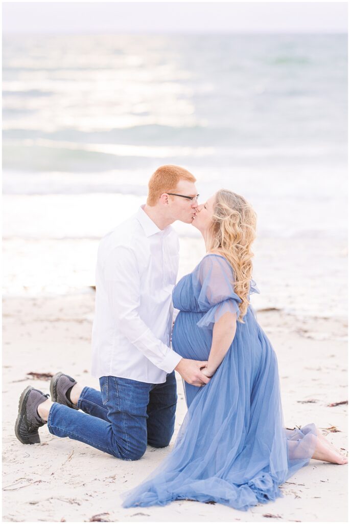 Man and woman kneeling in the sand and kissing while holding baby bump Cape Cod maternity photos