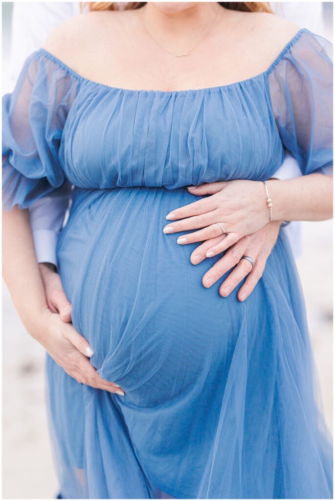 Close up of a man and woman's hands holding her baby bump as she wears a blue flowy dress