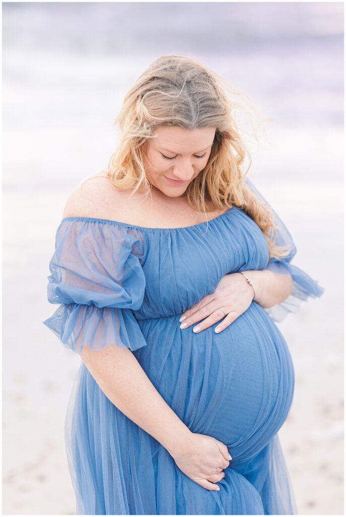 Woman holding and looking down at her baby bump wearing flow blue dress for maternity photos