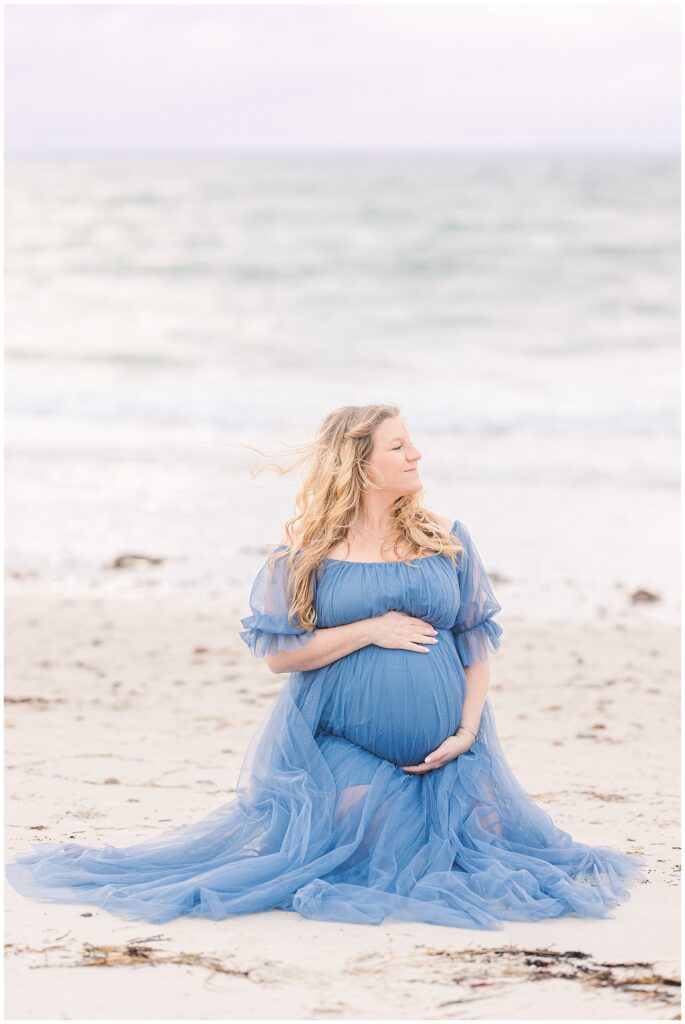Woman sitting on the beach as her hair blows in the breeze while holding her baby bump.
