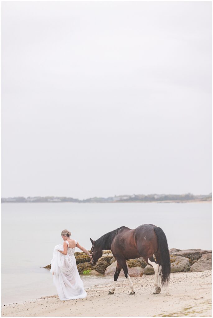 Bride holding her dress and leading her horse towards the ocean at Old Silver Beach in Falmouth, MA Cape Cod