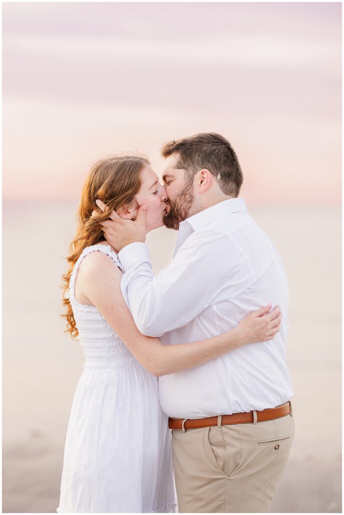 A man holding a woman's face as they kiss during their Gloucester beach engagement photos