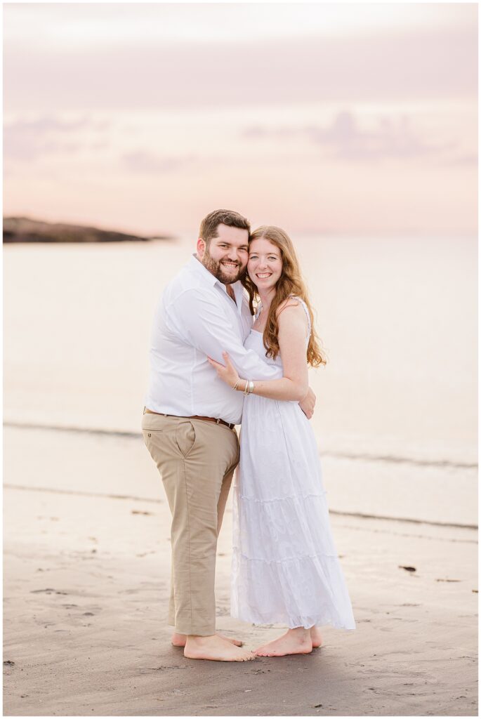 A couple hugging and smiling as they stand in front of waves at Wingaersheek Beach