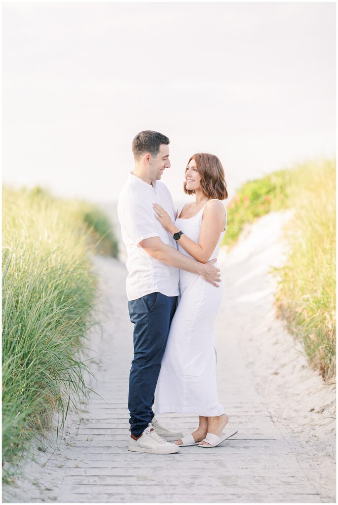 A couple stands close together on a sandy path lined with tall grass. They are both smiling and looking into each other's eyes.