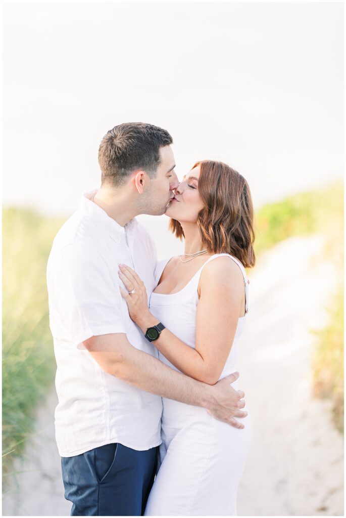 A couple is kissing on the sandy path. The man has his hand on the woman's waist, and she has her hand on his shoulder