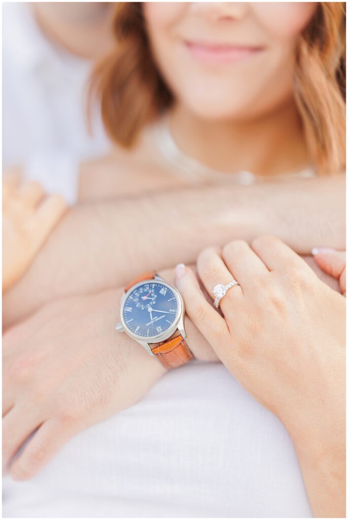 A close-up of the woman's hand resting on the man's arm. She is wearing a diamond engagement ring and a watch with a brown leather strap is visible on his wrist.