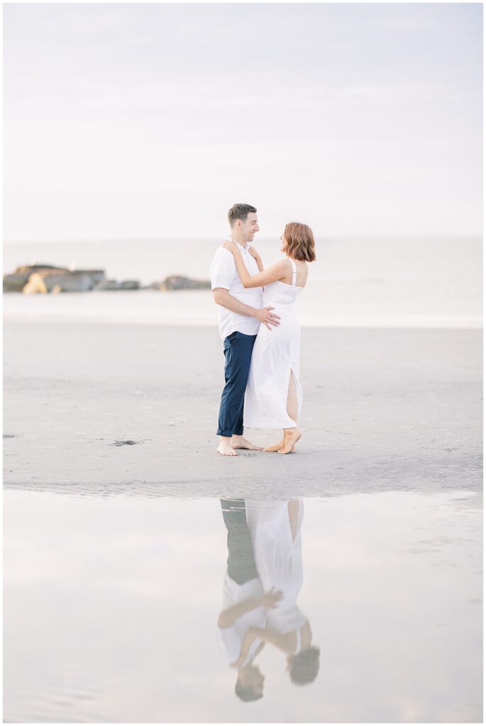 The couple stands on a beach with rocks and greenery in the background. They are looking into each other's eyes, smiling, and holding each other close