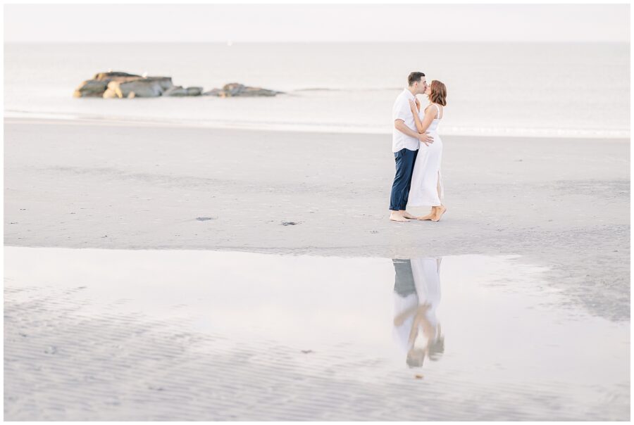 The couple is standing on a beach near the water. They are barefoot and embracing, with their reflection visible in a shallow pool of water on the sand