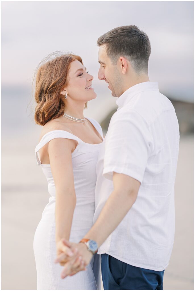 The couple is standing on the beach, holding hands and facing each other. They are smiling and looking into each other's eyes