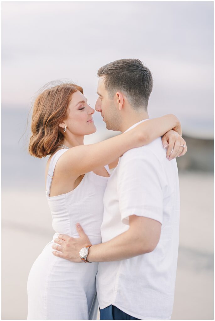 The couple is embracing on the beach with the ocean in the background. They are smiling at each other, and the woman has her arms around the man's neck