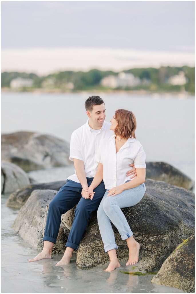 A couple sits on large rocks at a Gloucester beach, smiling at each other while holding hands with their feet in the water