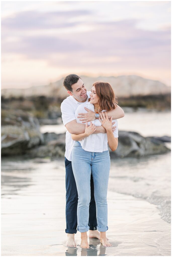 A man lifts a woman as they kiss, standing in the shallow water at a beach.