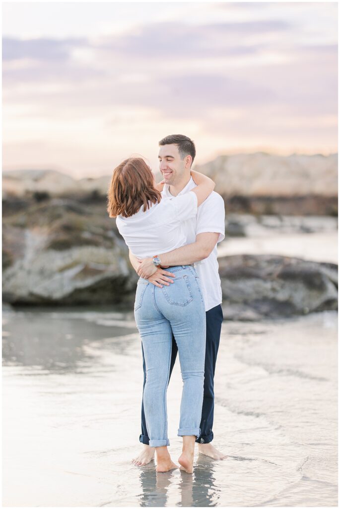 A couple embraces while standing barefoot in the water at Gloucester beach, with the man wrapping his arms around the woman.