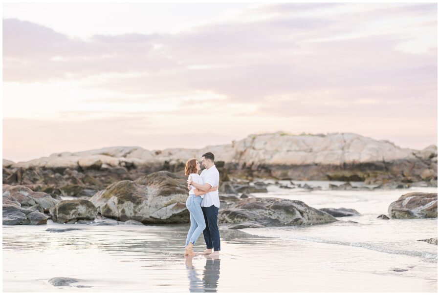 A couple hugs while standing in shallow water at Wingaersheek beach, surrounded by large rocks and a sunset sky.