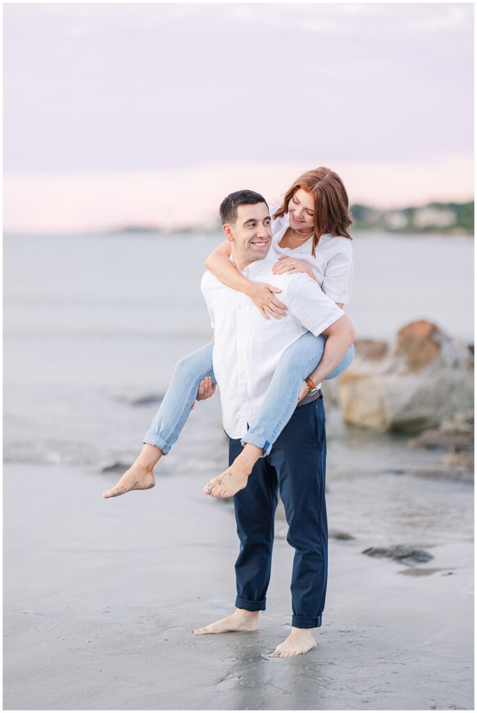 A man gives a woman a piggyback ride at a Gloucester beach, both laughing and barefoot on the sand near the water.