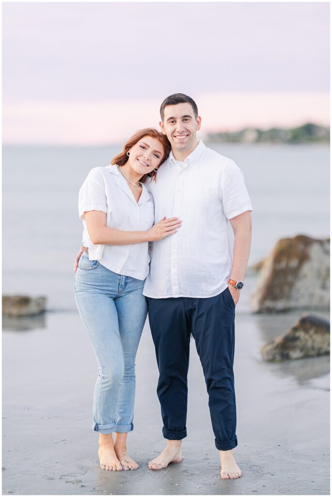 A couple stands side by side, smiling at the camera with their arms around each other and barefoot.