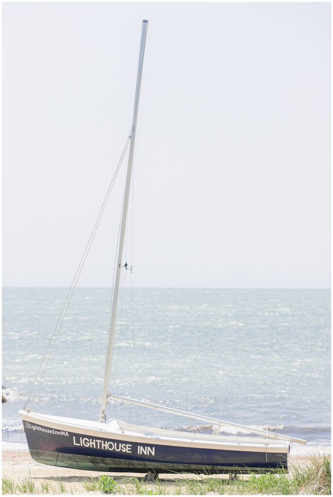 A small sailboat with “Lighthouse Inn” written on the side, resting on a sandy beach with the ocean in the background. Location: Lighthouse Inn, Cape Cod.