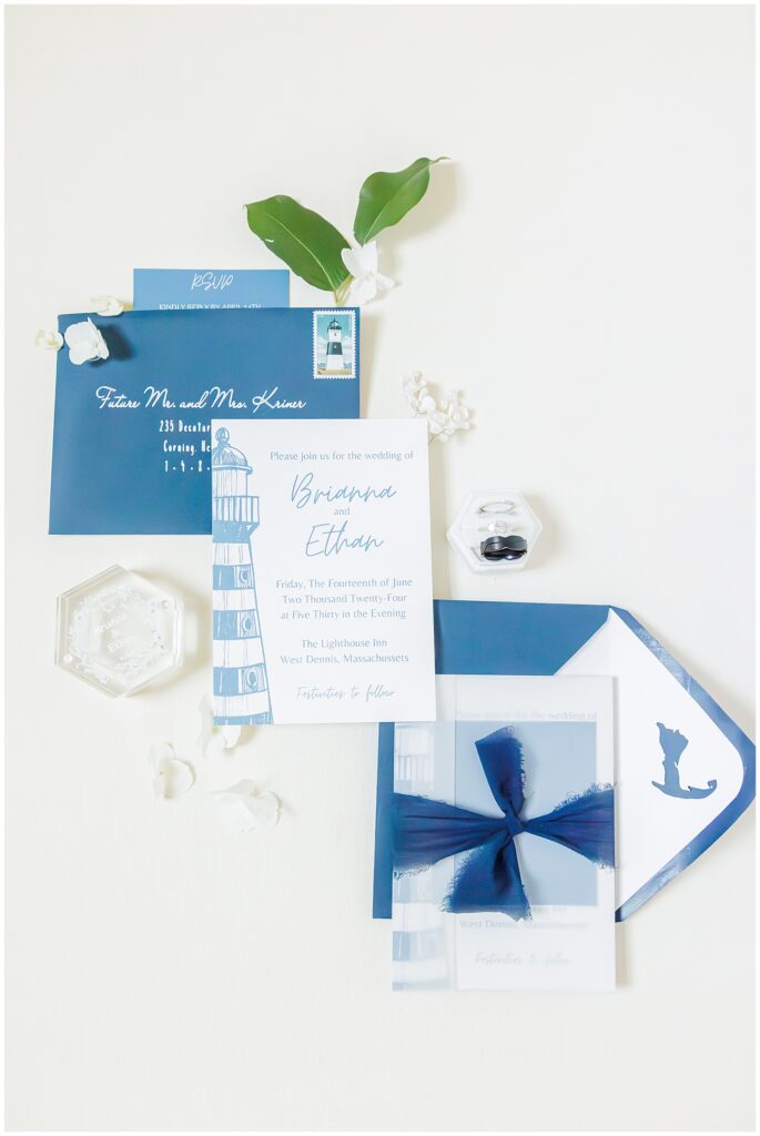 A wedding invitation set featuring blue and white stationery, a small box with a ring, and green leaves for decoration. The invitation mentions “The Lighthouse Inn, West Dennis, Massachusetts.”
