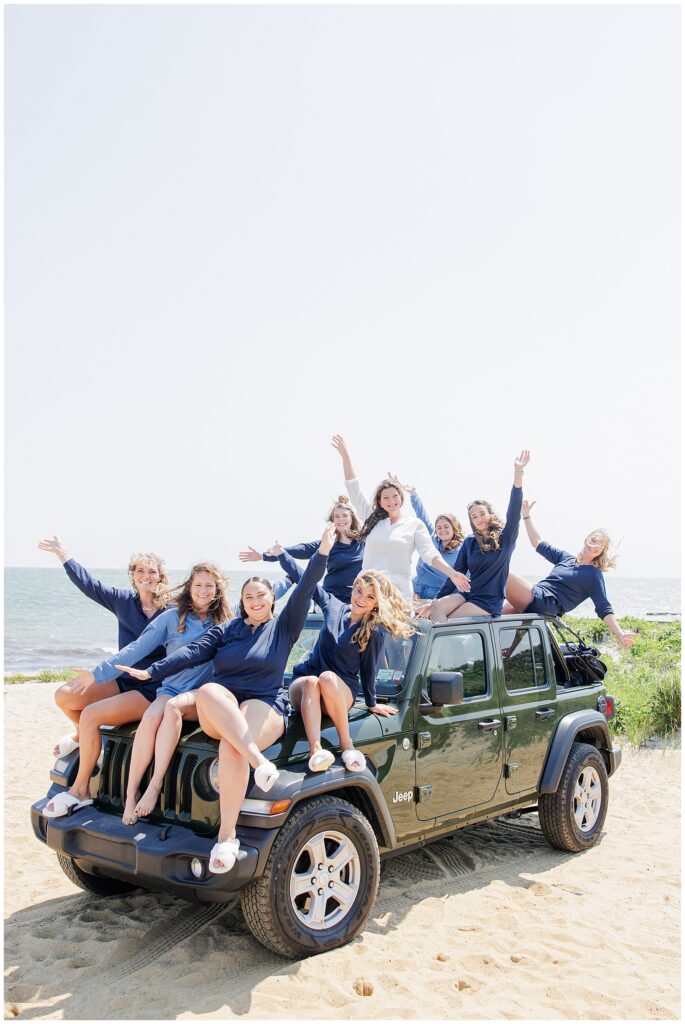 A group of women sitting and standing on a green Jeep on the beach, all wearing blue shirts and smiling with arms raised.