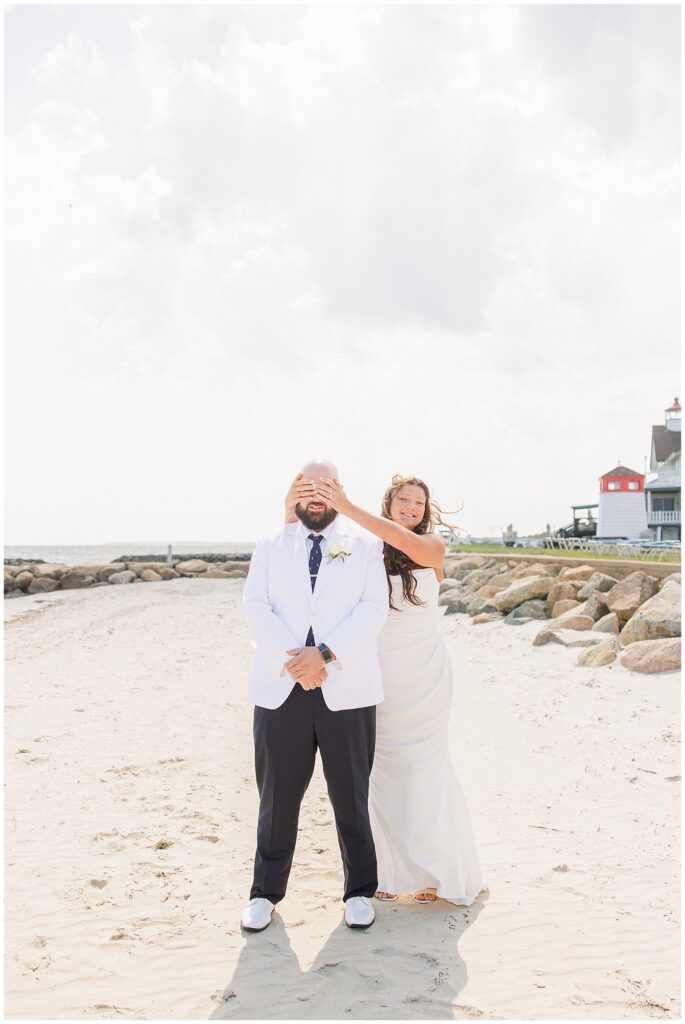 A bride in a white dress covering the eyes of a groom in a white jacket and navy pants on a sandy beach, with a rocky shore and a lighthouse in the background. Location: Lighthouse Inn, Cape Cod.