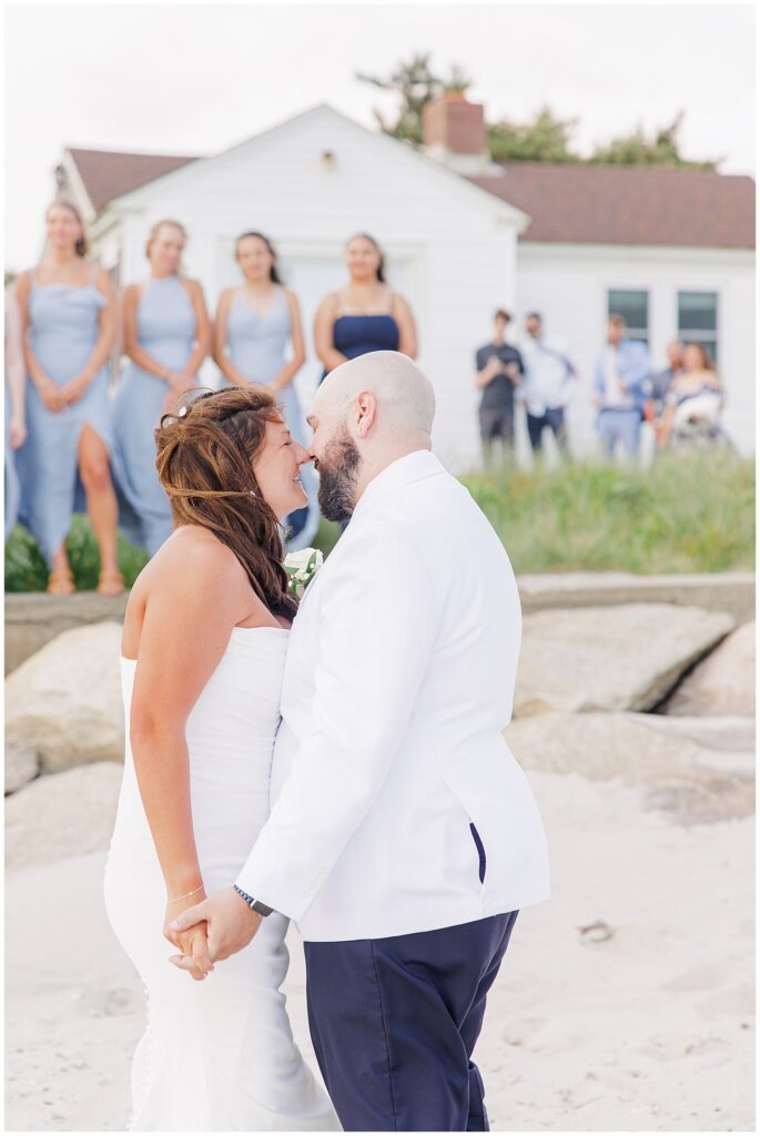 A bride and groom holding hands and touching foreheads on a beach, with bridesmaids in light blue dresses standing in the background near a white building.