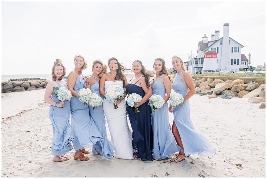 A bride in a white dress and six bridesmaids in light blue and navy dresses posing on a sandy beach, each holding a bouquet of white and blue flowers. Location: Lighthouse Inn, Cape Cod.