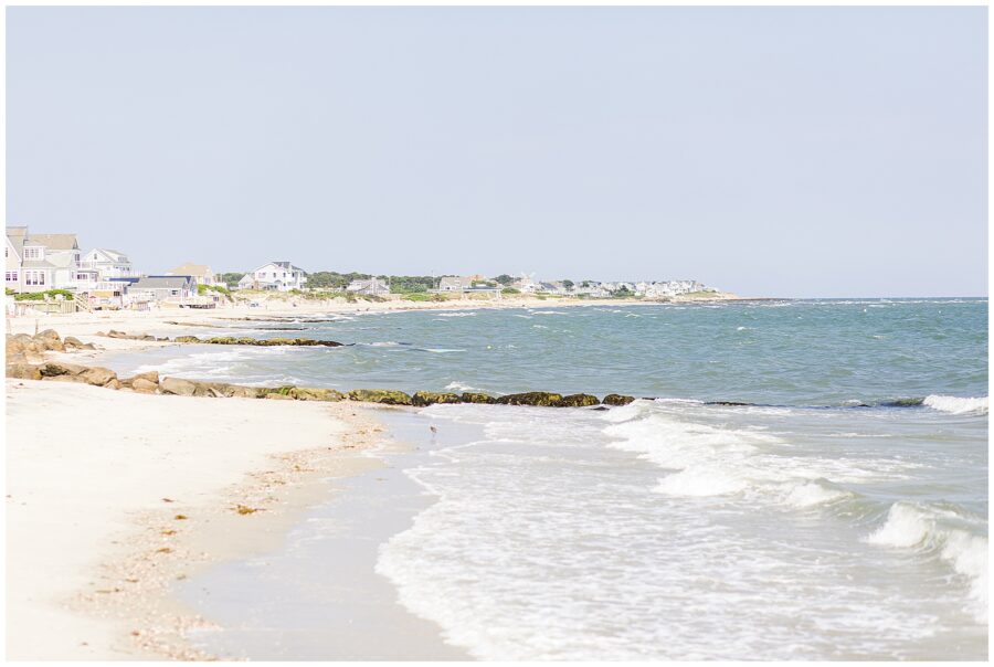 View of a sandy beach with gentle waves, beach houses, and greenery in the distance.