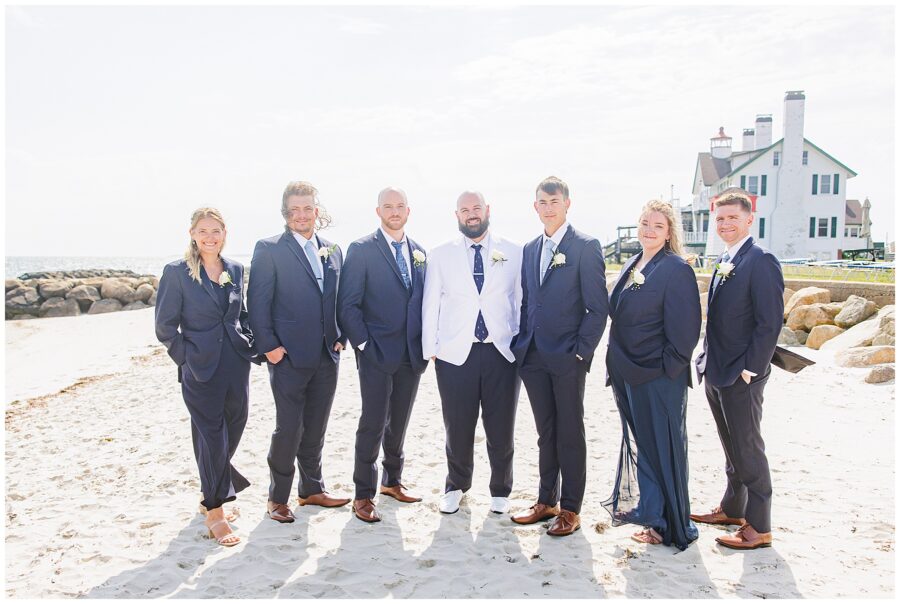 The groom and six groomsmen standing on the beach, wearing navy blue suits, with the groom in a white jacket. A lighthouse and building are visible in the background. Location: Lighthouse Inn, Cape Cod.