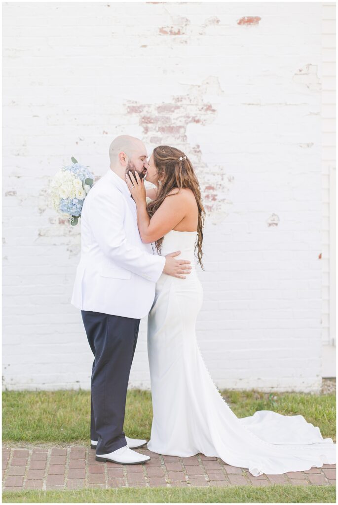 A bride and groom kissing in front of a white brick wall with faded red paint. The bride is holding a bouquet of white and blue flowers.