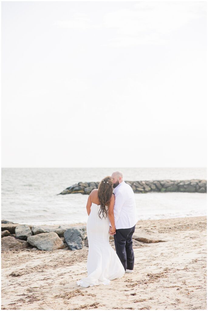 A bride and groom walking hand in hand on a sandy beach, with the groom kissing the bride’s head. Rocks and the ocean are in the background.