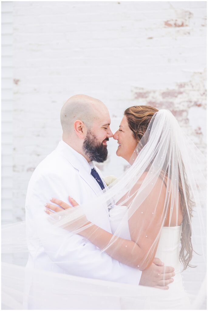 Close-up of a bride and groom facing each other and smiling, the bride’s veil draped around them, standing in front of a white brick wall.