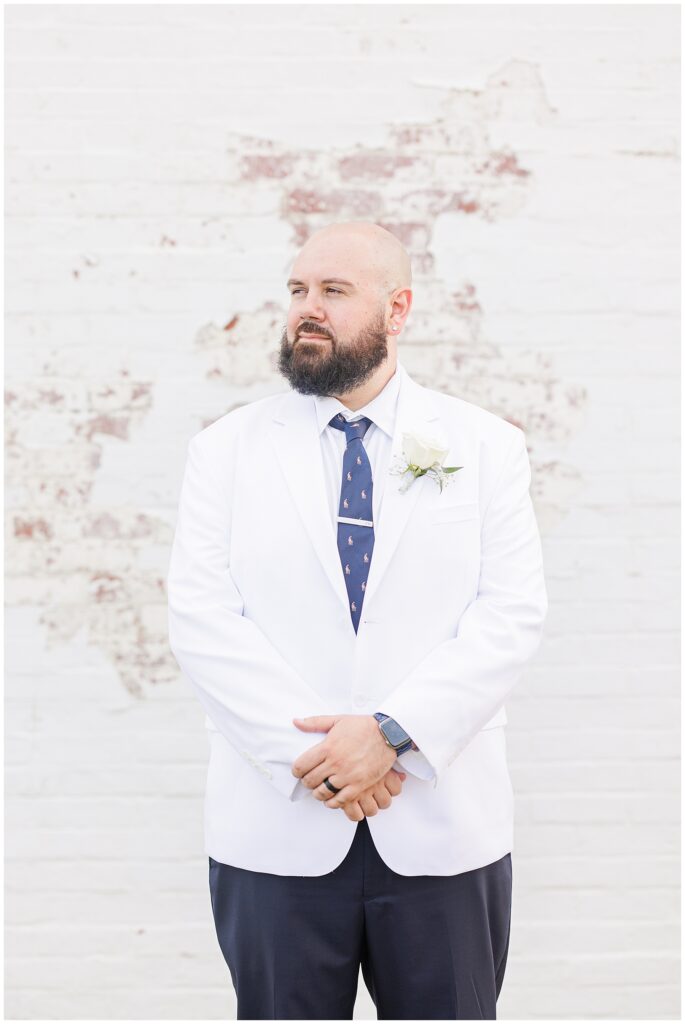 The groom, dressed in a white jacket and navy pants, standing with hands clasped in front of a white brick wall.