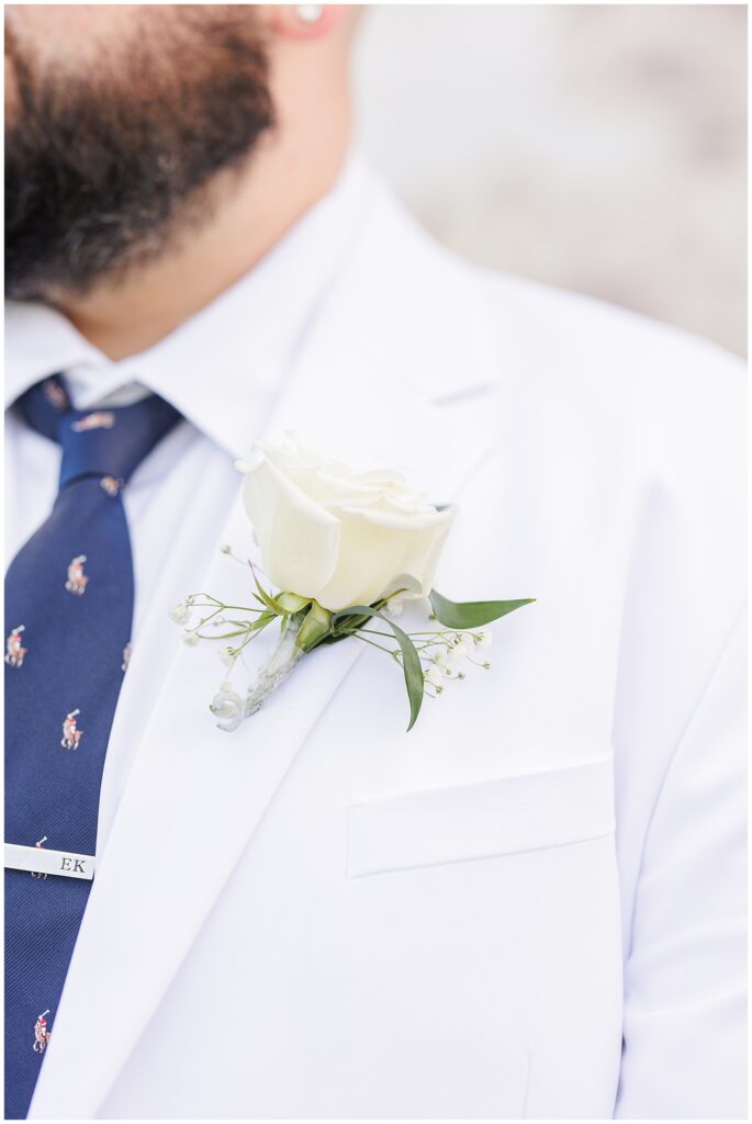 Close-up of the groom’s boutonnière, a white rose pinned to his white jacket lapel.