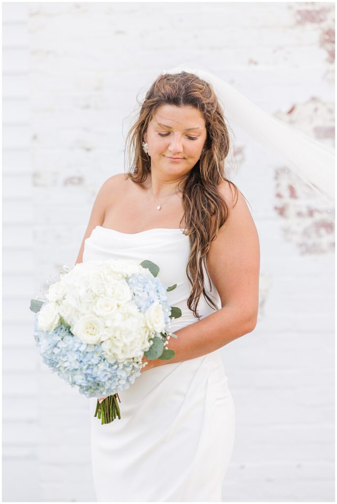 The bride holding a bouquet of white roses and light blue hydrangeas, standing in front of a white brick wall. Location: Lighthouse Inn, Cape Cod.