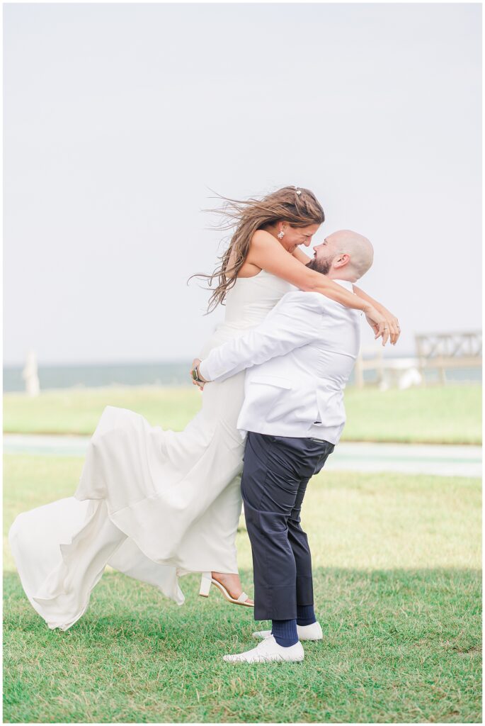 The groom lifting the bride in the air as they both smile, standing on a grassy area near the ocean.