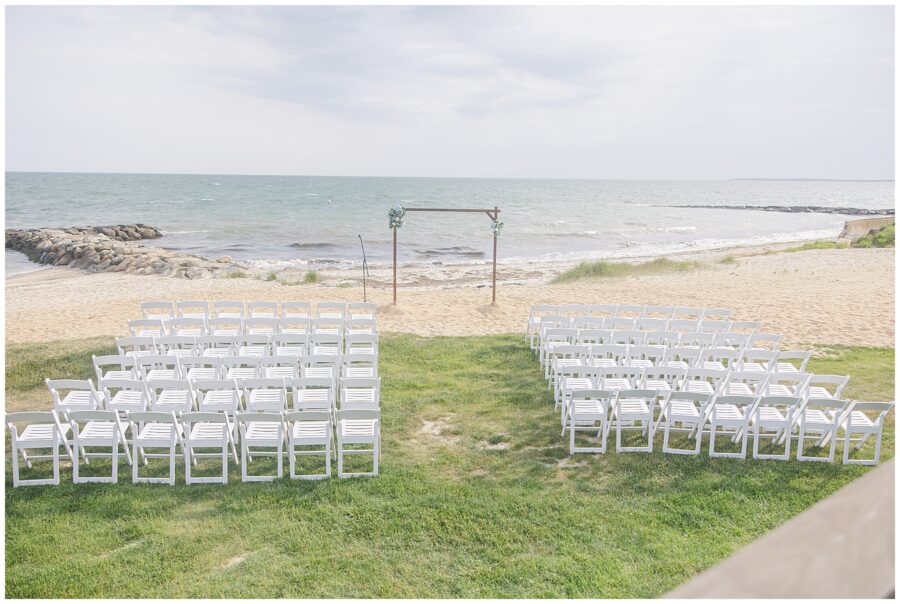 A wedding ceremony setup on a grassy area by the beach, with rows of white chairs facing an arch decorated with flowers. Location: Lighthouse Inn, Cape Cod.