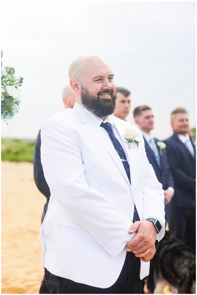 The groom, wearing a white jacket, smiling and standing with his groomsmen on the beach.