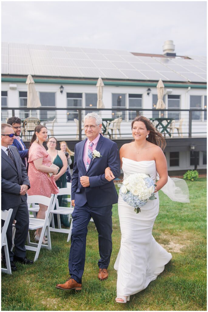 The bride, holding a bouquet of white roses and blue hydrangeas, walking down the aisle with her father.