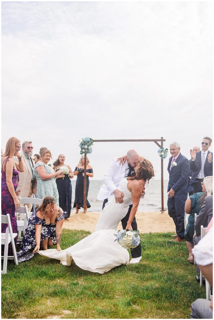 he groom dipping the bride for a kiss as guests cheer and one adjusts the bride’s dress. Location: Lighthouse Inn, Cape Cod.