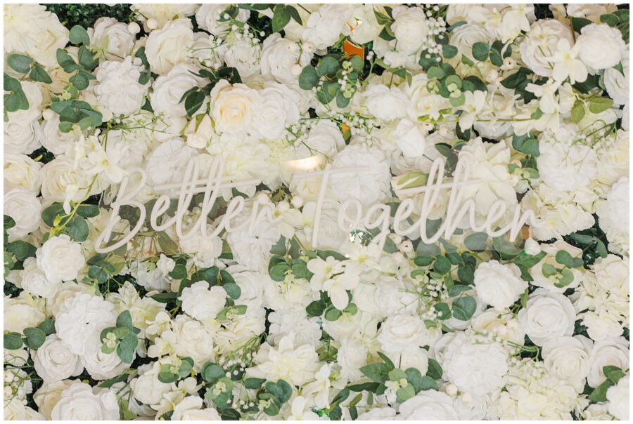 A decorative floral wall with white roses and green leaves, featuring a sign that says “Better Together.”