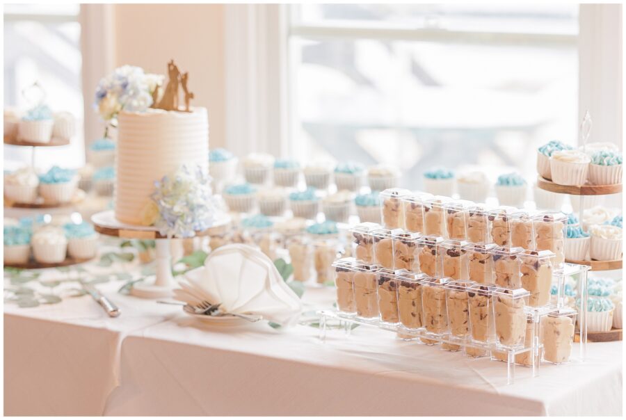 Dessert table featuring a white cake with a wooden bride and groom topper, surrounded by blue and white frosted cupcakes and dessert cups.