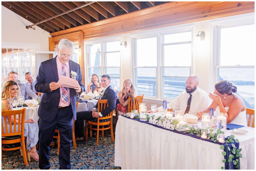 An older man giving a speech at a reception, while the bride and groom sit at a table nearby, with guests seated around them.