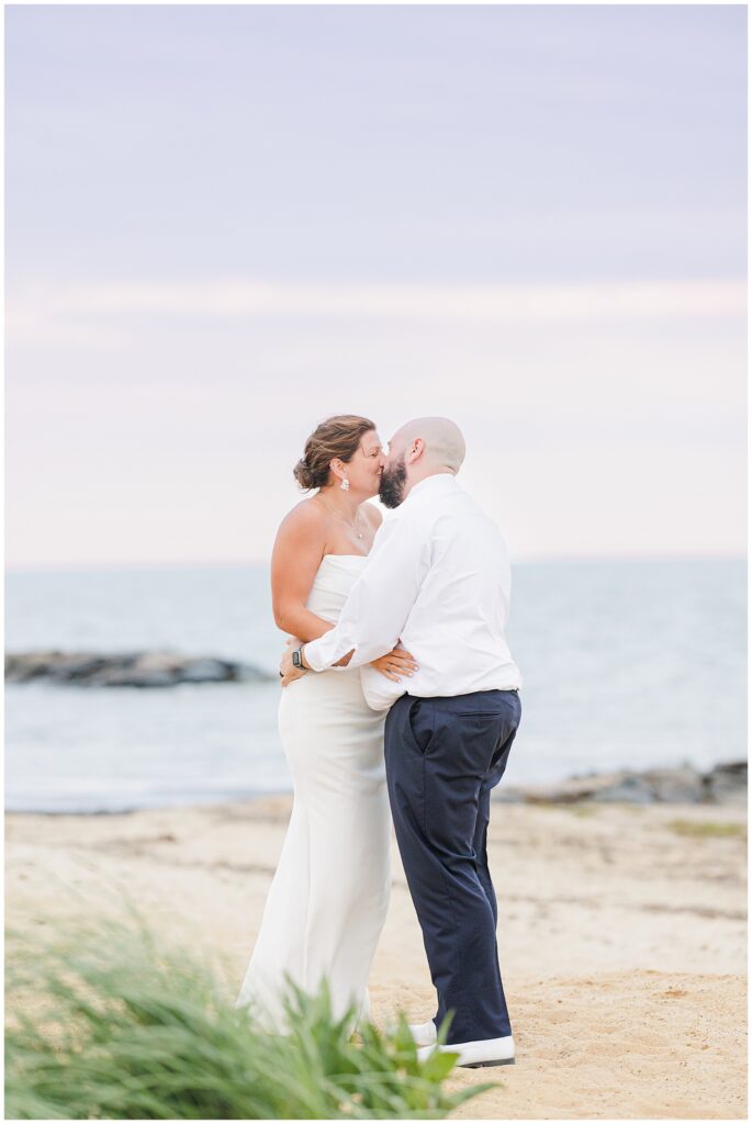 Bride and groom kissing on the beach with the ocean in the background.