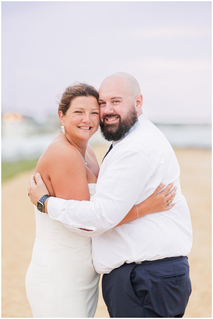 Bride and groom embracing and smiling at the camera on a sandy beach.