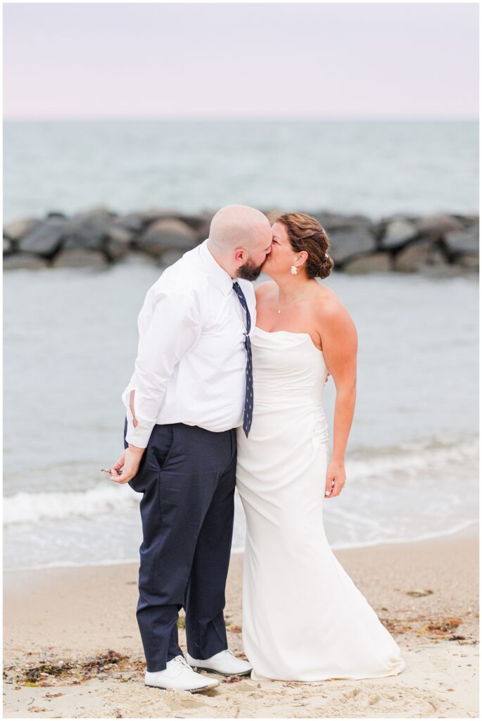 Bride and groom kissing on the beach, with the ocean and rocks in the background.