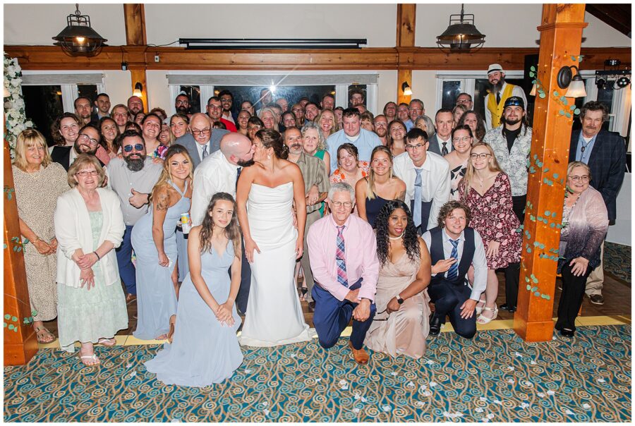 Large group photo of wedding guests with the bride and groom kissing in the center. Location: Lighthouse Inn, Cape Cod.